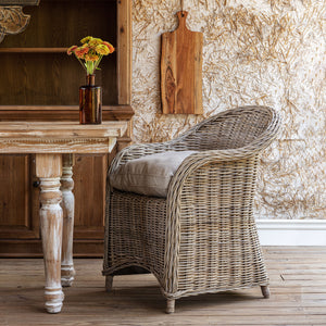 A woven wicker armchair with a light beige cushion sits beside a rustic wooden table in a styled interior setting with a textured wall and wooden decor.