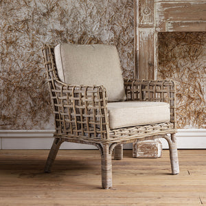 A rustic woven rattan armchair with thick beige fabric cushions, set against a distressed wood-paneled wall and light wood floor.