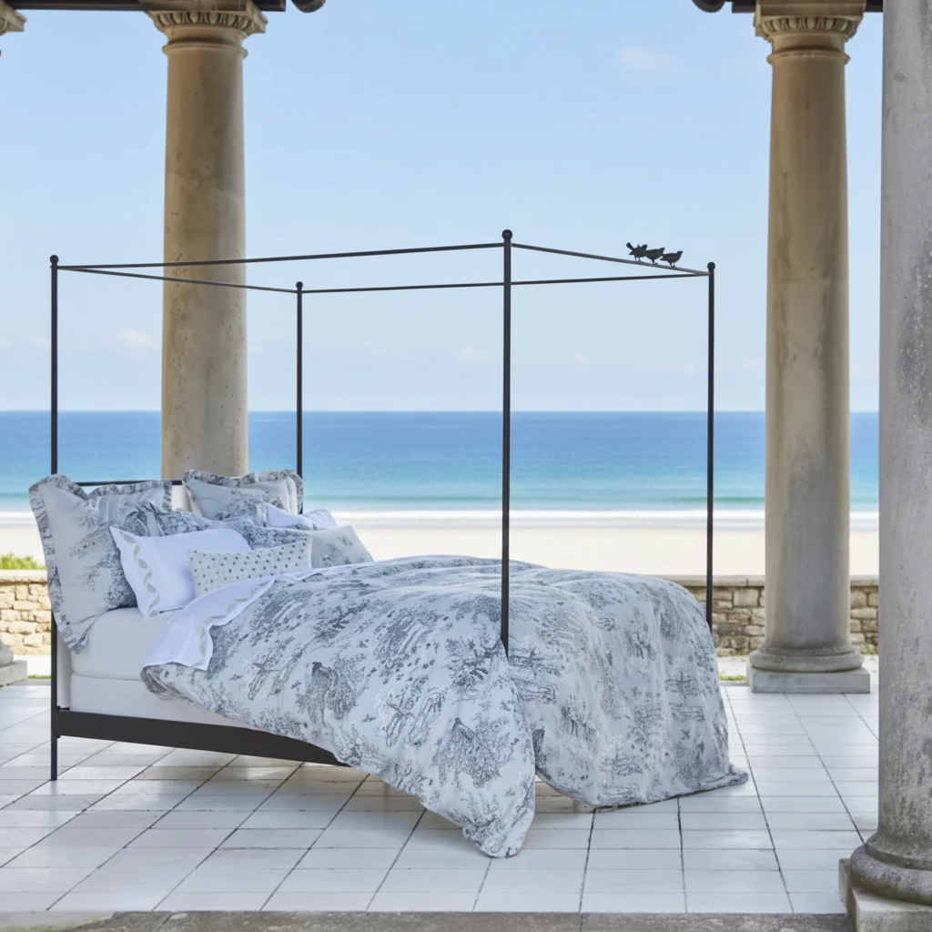 A black metal four-poster bed with a gray and white toile patterned duvet and matching pillows, styled on a tiled porch with a beach and ocean view in the background.