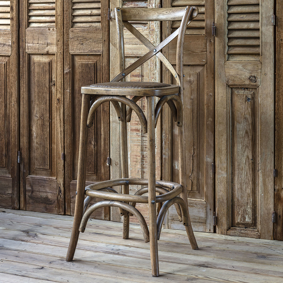 A rustic wooden bar stool with a bentwood frame, woven seat, and crossed backrest, set against a matching weathered wooden door and plank floor.