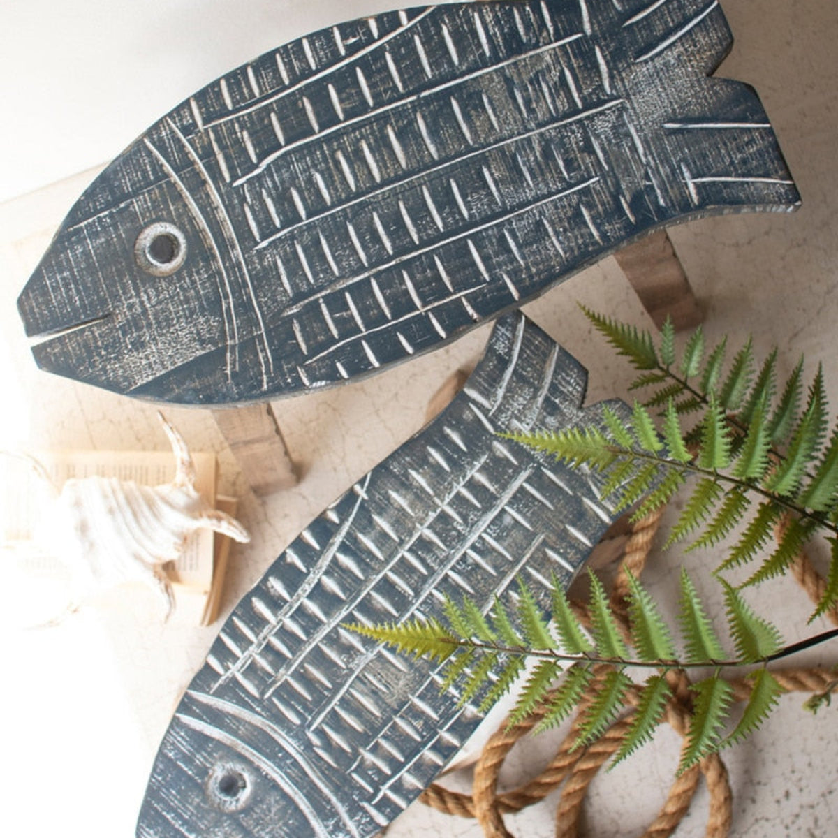 Two fish-shaped decorative wall pieces made from distressed dark wood, arranged on a light surface with a fern leaf and rope as styling props.