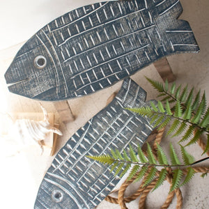 Two fish-shaped decorative wall pieces made from distressed dark wood, arranged on a light surface with a fern leaf and rope as styling props.