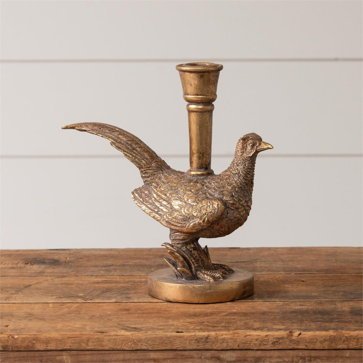 A bronze-finished pheasant figurine candlestick holder with a white candle inserted, displayed on a wooden surface with autumn leaves against a white paneled wall.