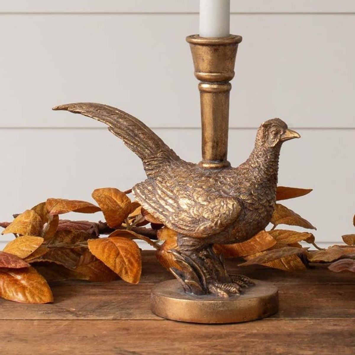 A bronze-finished pheasant figurine candlestick holder with a white candle inserted, displayed on a wooden surface with autumn leaves against a white paneled wall.