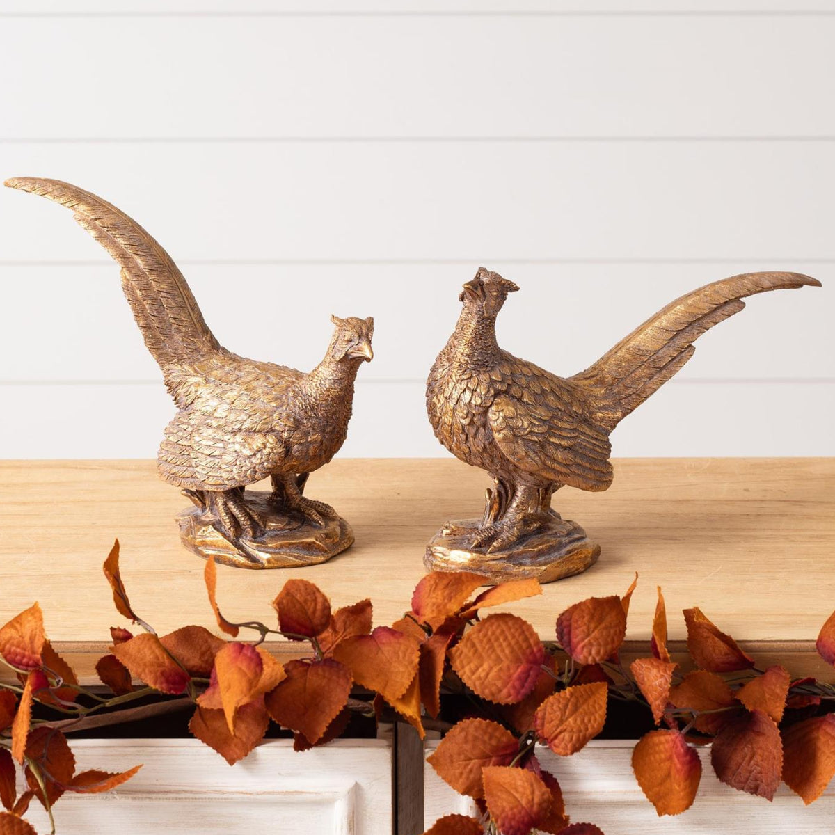 Two metallic pheasant figurines with a weathered bronze finish, displayed on a dining table runner surrounded by autumn leaves, pumpkins, and candles in a warm, decorated dining room.