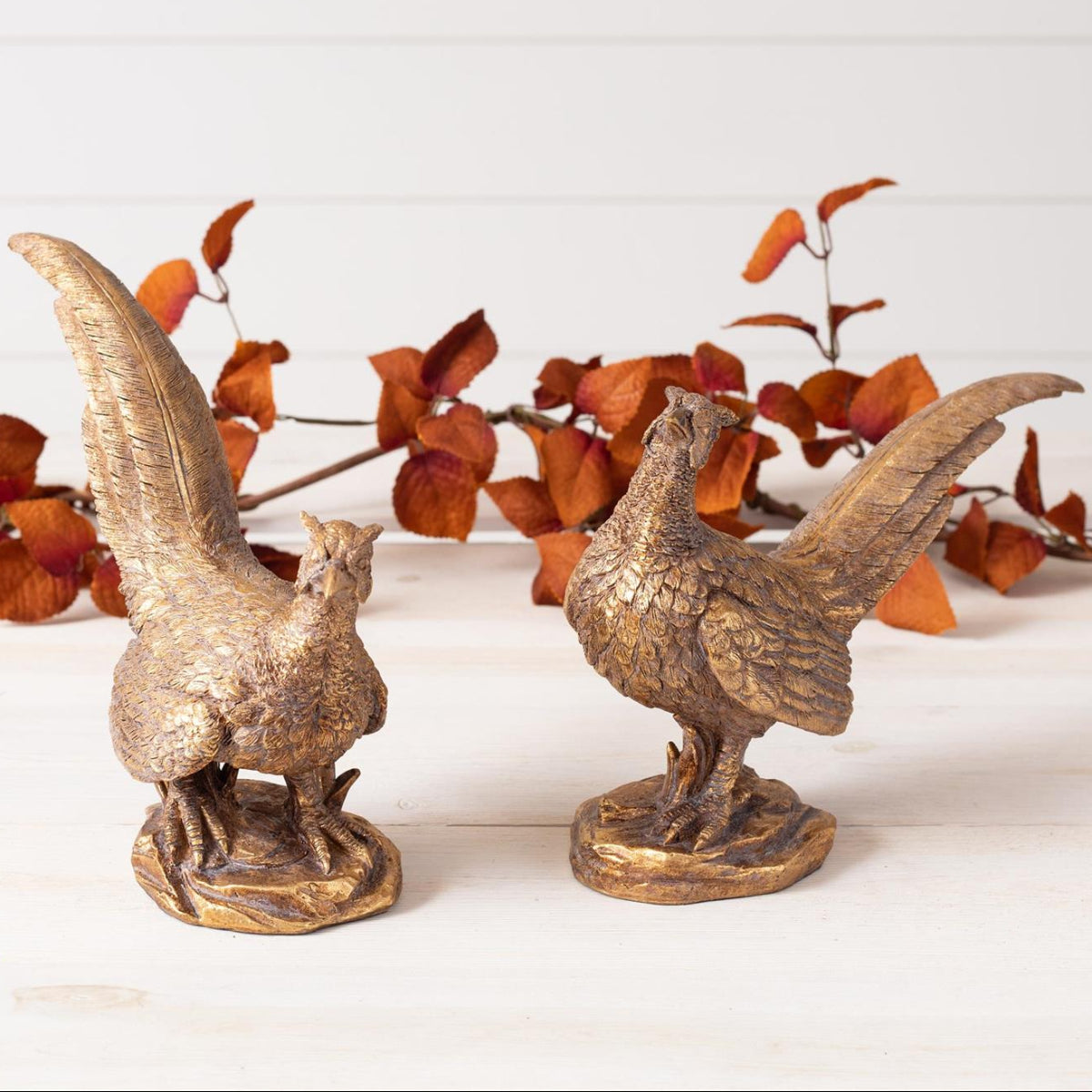 Two decorative pheasant figurines with a metallic gold finish, displayed on a white wooden surface with dried orange foliage in the background.