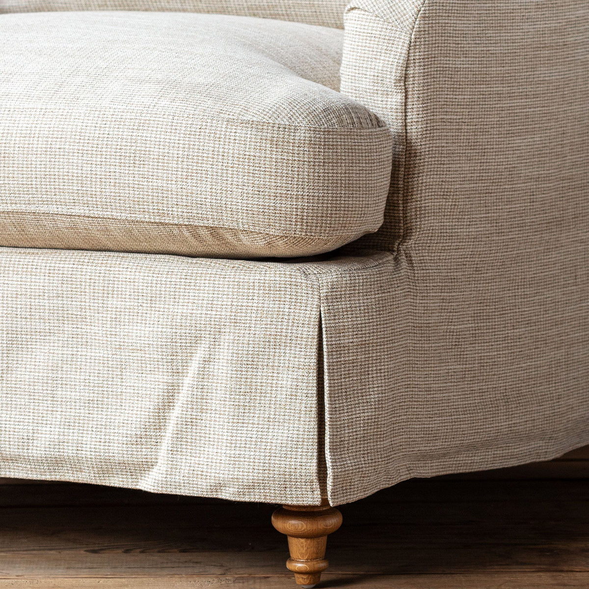 Close-up of a beige textured fabric slipcover on an upholstered piece of furniture, showing the seat cushion, armrest, skirted base, and turned wooden legs on a dark wood floor.