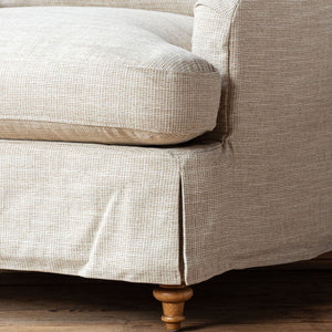 Close-up of a beige textured fabric slipcover on an upholstered piece of furniture, showing the seat cushion, armrest, skirted base, and turned wooden legs on a dark wood floor.