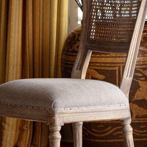 A close-up of a rustic dining chair with a light beige upholstered seat and a woven cane backrest, set against a warm, patterned background with curtains and a decorative vase.