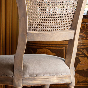 A close-up of a vintage-style wooden dining chair with a woven cane backrest and a light beige upholstered seat cushion, set against a warm, patterned background.