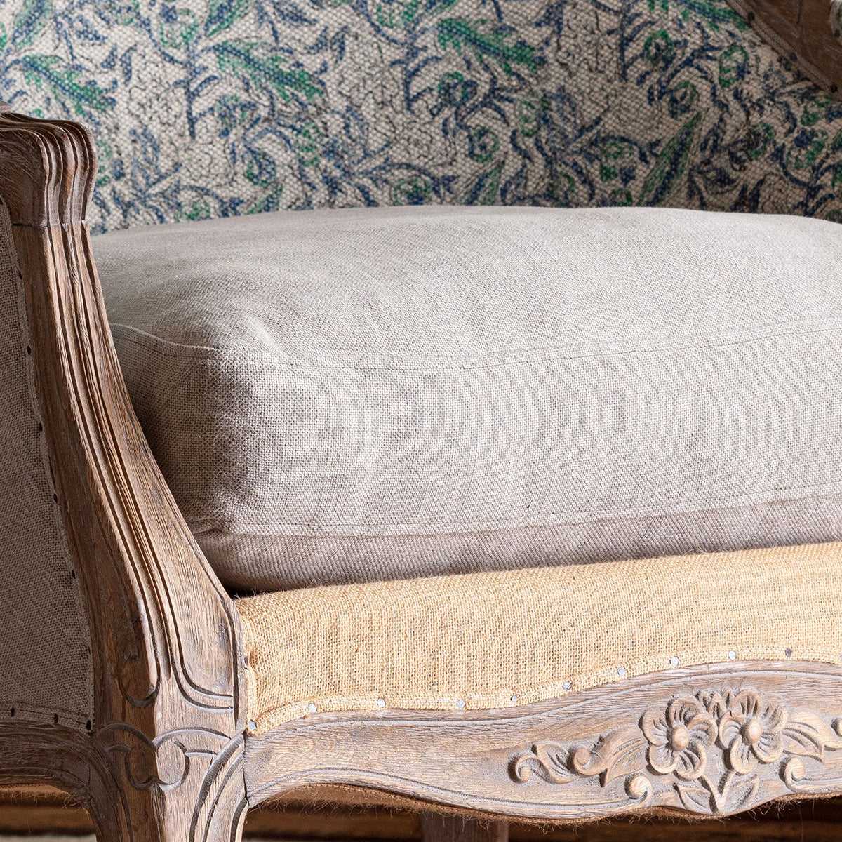A close-up of a light beige upholstered cushion resting on a carved wooden armchair with woven jute seat panels and a patterned green and blue fabric backrest.