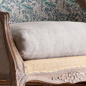 A close-up of a light beige upholstered cushion resting on a carved wooden armchair with woven jute seat panels and a patterned green and blue fabric backrest.