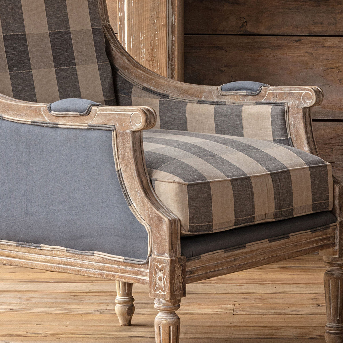 A classic French-style accent chair with a distressed light brown wood frame, featuring striped beige and gray plaid upholstery on the backrest and seat, and solid gray fabric on the armrests, set against a rustic wood-paneled wall and floor.