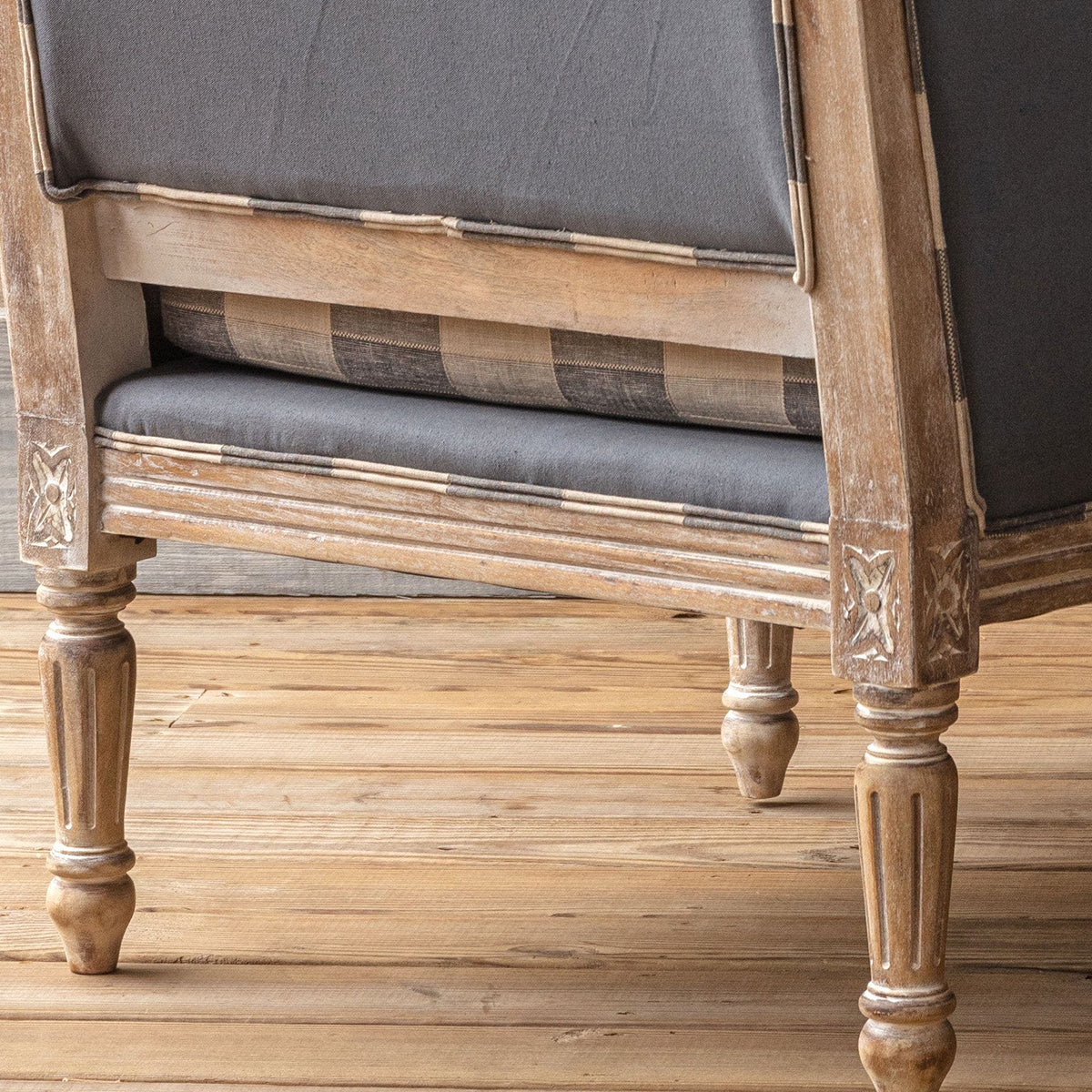 Close-up of a rustic wooden armchair with dark gray upholstered cushions and carved turned legs, shown on a light wood floor.