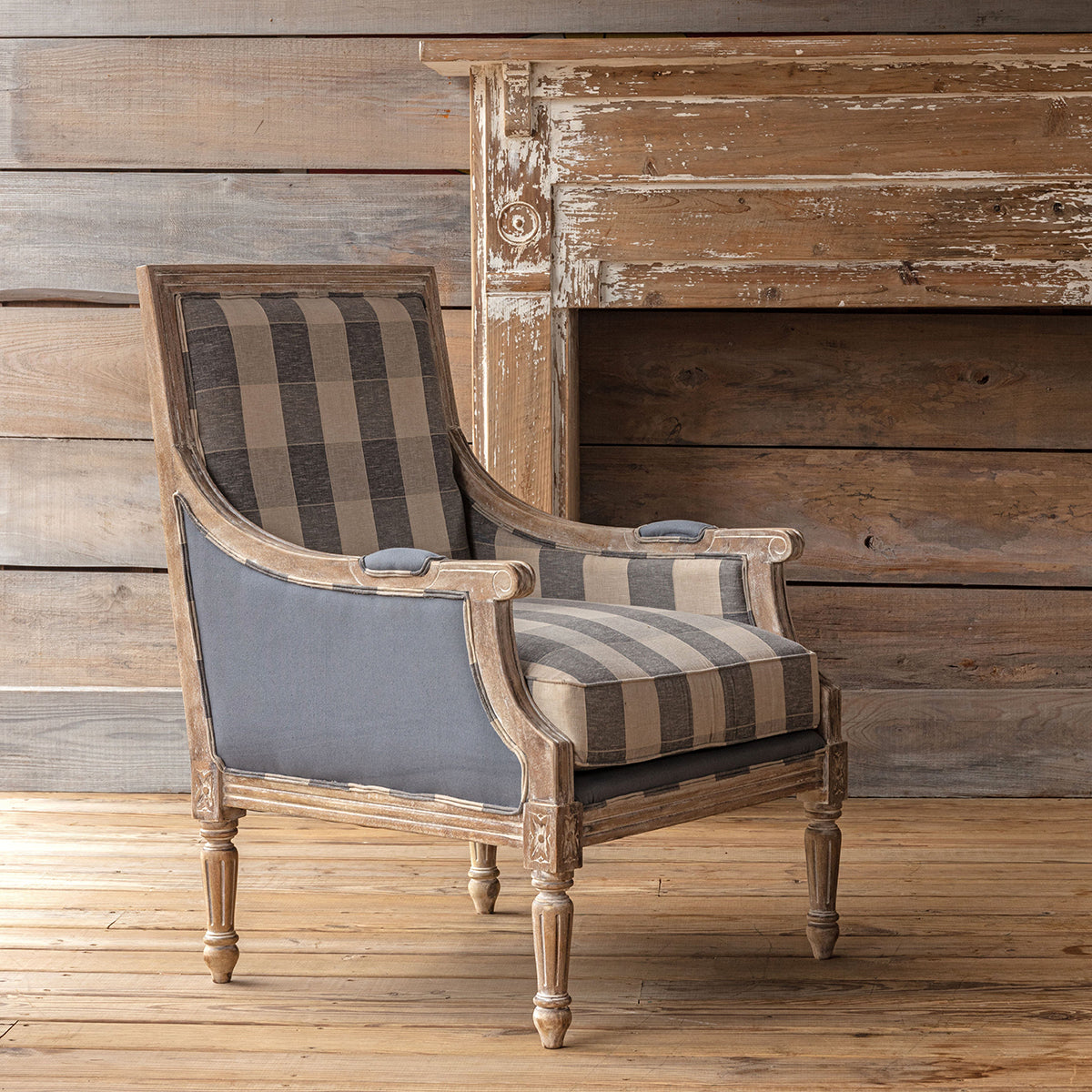A classic French-style accent chair with a distressed light brown wood frame, featuring striped beige and gray plaid upholstery on the backrest and seat, and solid gray fabric on the armrests, set against a rustic wood-paneled wall and floor.