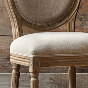 A close-up of a rustic wooden dining chair with a distressed finish, featuring a round upholstered backrest and a light beige textured fabric seat cushion.