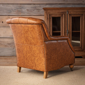 A classic brown leather armchair with nailhead trim, shown from a three-quarter angle in a rustic room setting with a wooden cabinet and paneled wall.