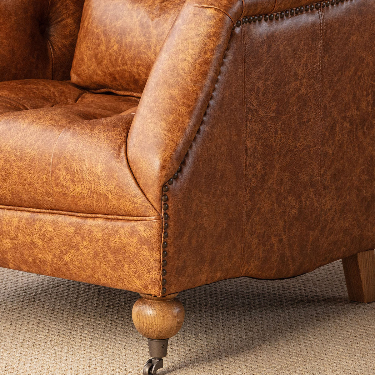 Close-up of a brown distressed leather armchair with button tufting, nailhead trim, and turned wooden legs on a textured beige carpet.