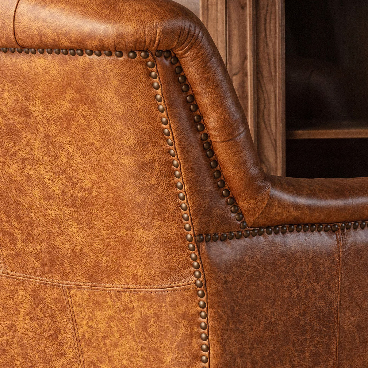 Close-up of a rustic brown leather armchair showing the textured grain, curved armrest, and dark metal nailhead trim.