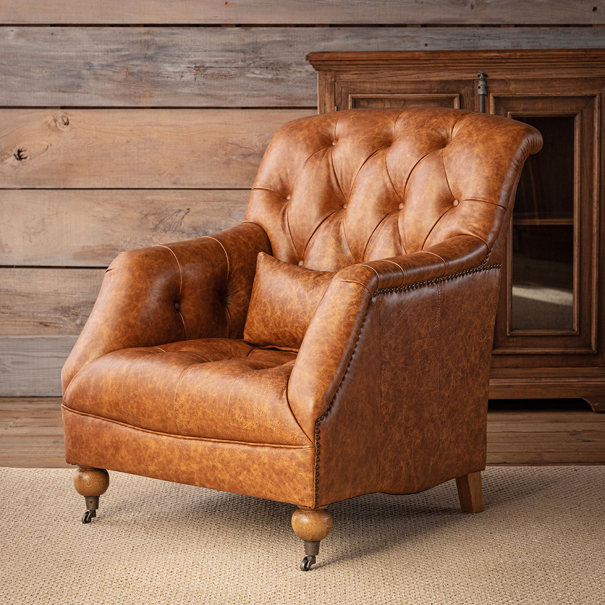 A classic tufted leather armchair in a warm cognac brown, styled in a rustic room setting with a wooden paneled wall and a dark wood cabinet in the background.