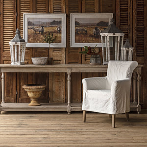 A rustic styled interior scene featuring a white slipcovered armchair in front of a wooden console table, with two framed landscape prints, lanterns, and a decorative urn against a wooden shutter wall.