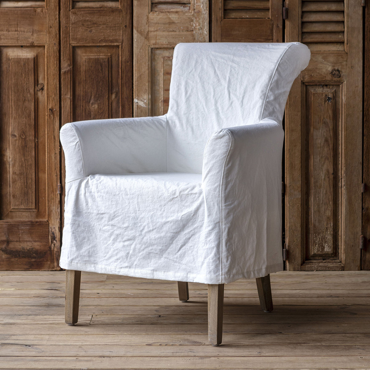A white fabric slipcover fitted over an armchair, shown in front of rustic wooden doors and on a wooden floor.
