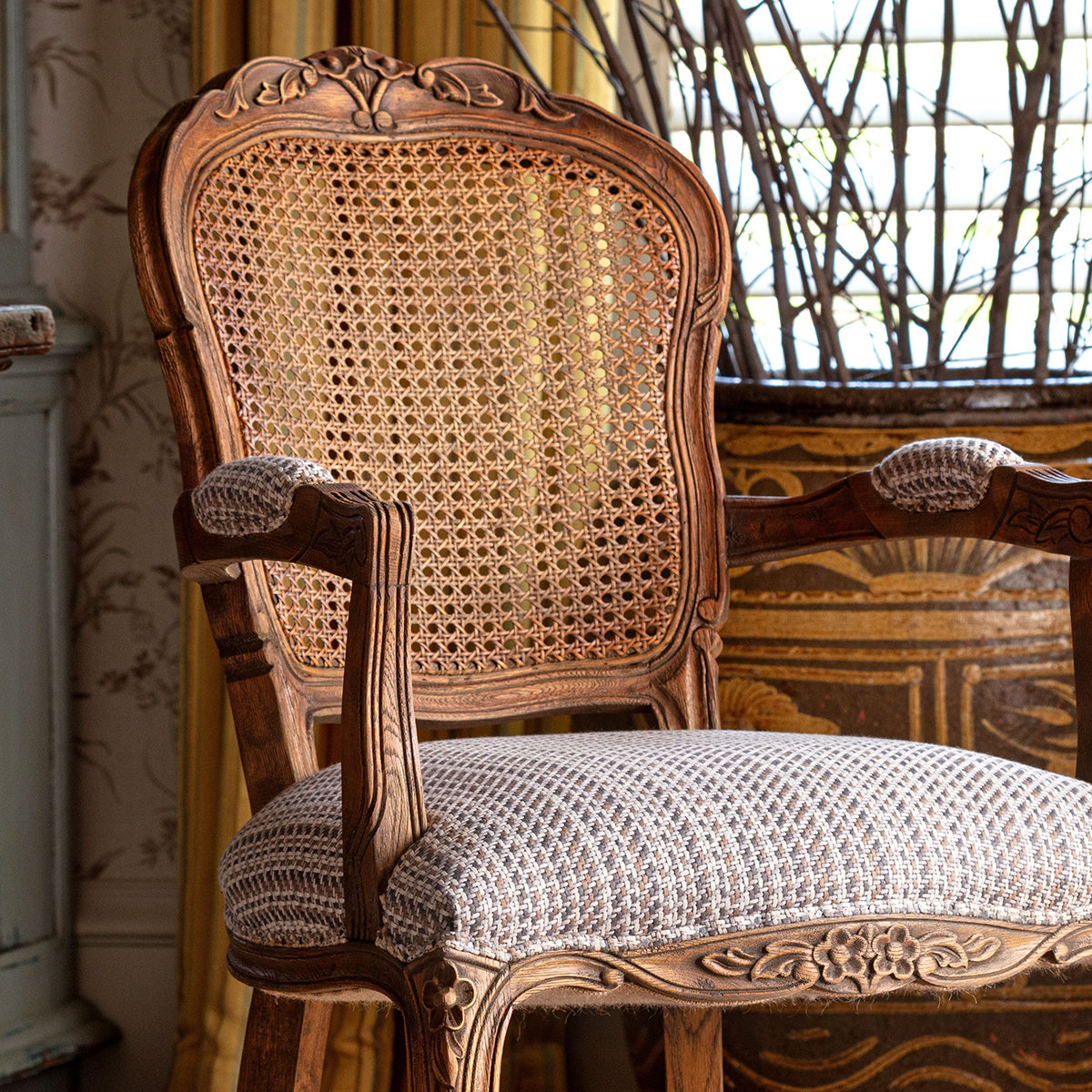 An ornate vintage-style wooden armchair with a woven cane backrest and textured cream upholstery, set in a decorated interior room.