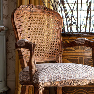 An ornate vintage-style wooden armchair with a woven cane backrest and textured cream upholstery, set in a decorated interior room.
