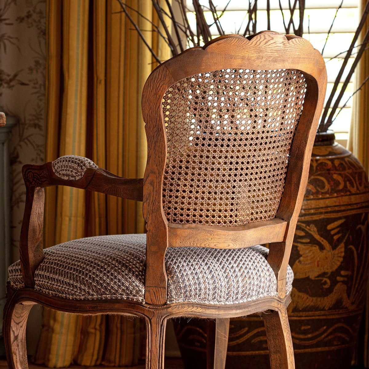 A classic French-style wooden armchair with a woven cane backrest and a textured gray and white patterned seat cushion, set in a warm, decorated room with yellow curtains and a large decorative vase.