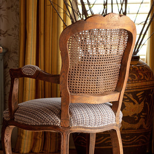 A classic French-style wooden armchair with a woven cane backrest and a textured gray and white patterned seat cushion, set in a warm, decorated room with yellow curtains and a large decorative vase.