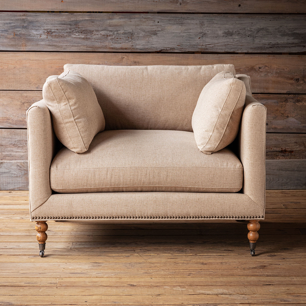 A beige upholstered armchair with two matching throw pillows, shown on a rustic wood plank floor against a matching wood-paneled wall.