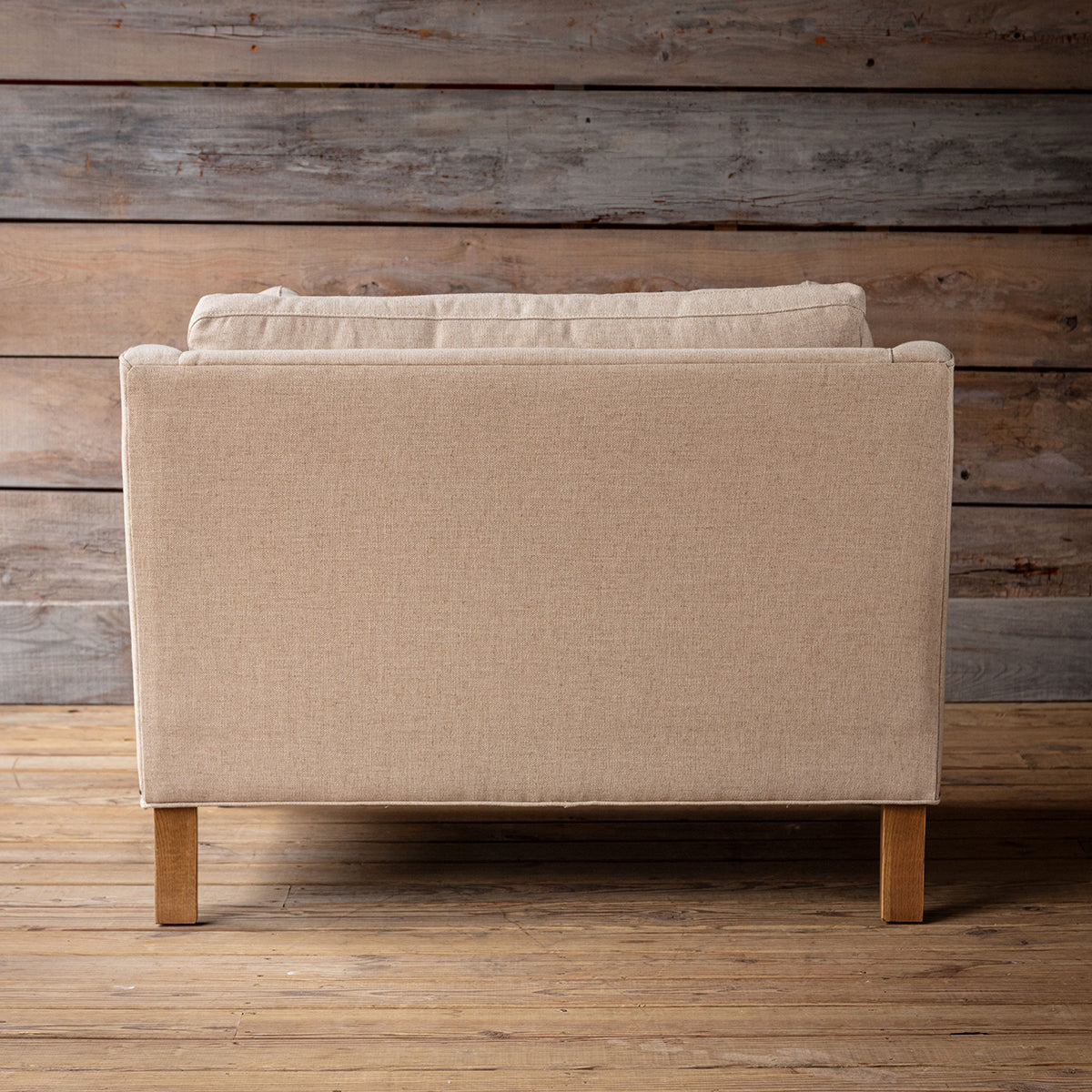 A beige upholstered armchair shown from the back, featuring a high back and light wood legs, set against a rustic wood plank wall and floor.