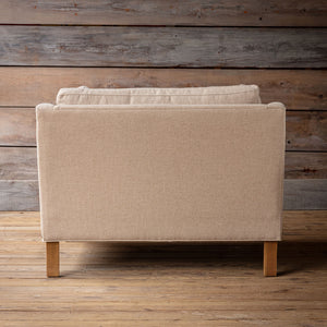A beige upholstered armchair shown from the back, featuring a high back and light wood legs, set against a rustic wood plank wall and floor.