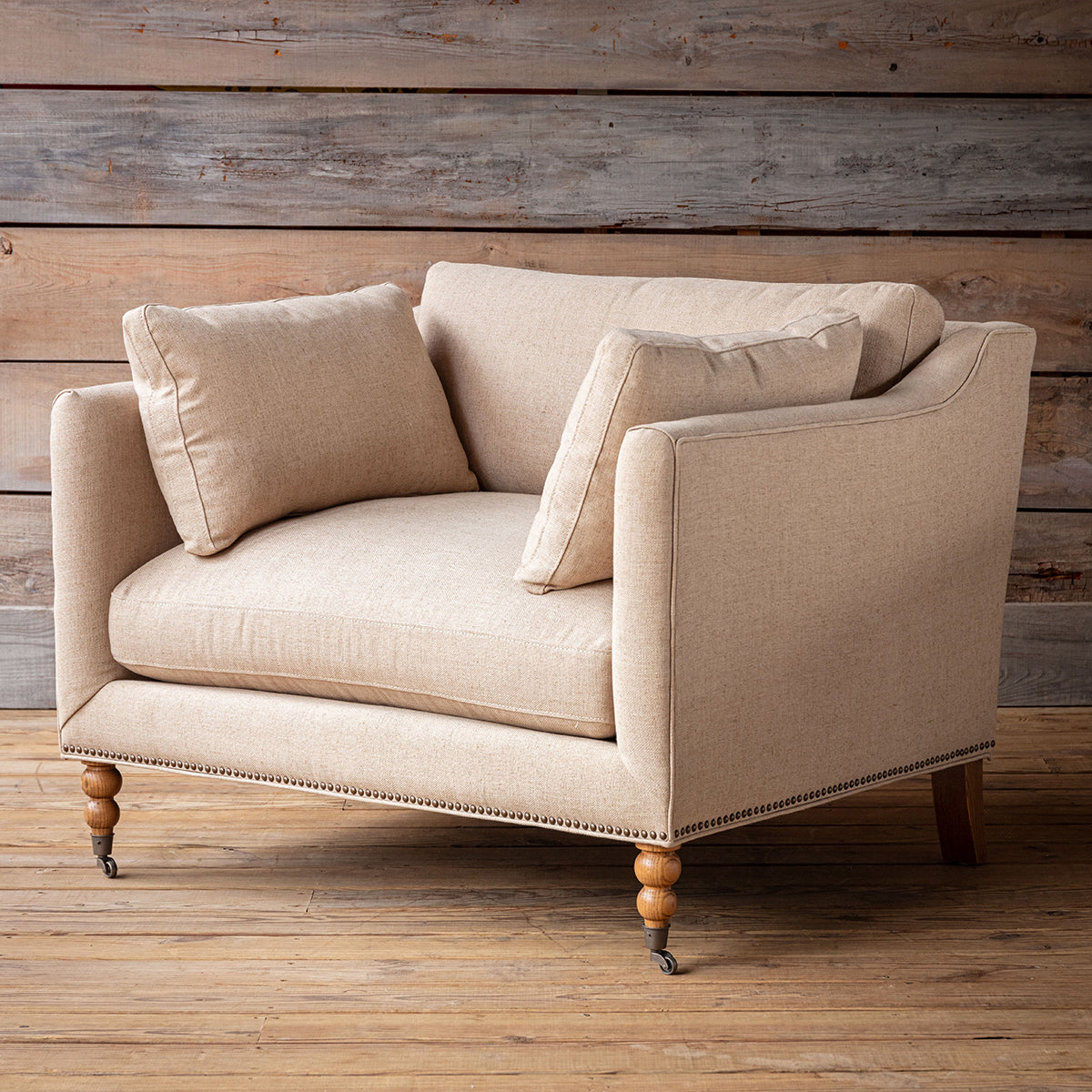 A beige upholstered armchair with two matching throw pillows, shown on a rustic wood plank floor against a matching wood-paneled wall.