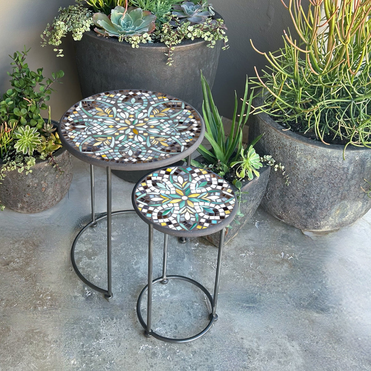 Two round nesting tables with Ojai Valley mosaic tops in various shades of green, blue, and beige, standing on iron or aluminum bases.