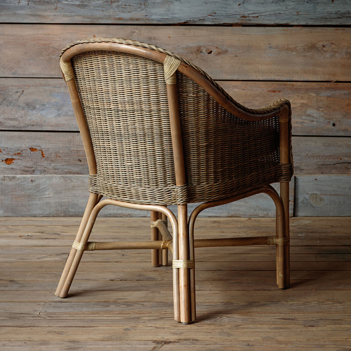 A natural woven rattan armchair with a curved back, rope-wrapped arm details, and a light beige cushion, set against a rustic wood plank wall and floor.