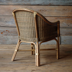 A natural woven rattan armchair with a curved backrest and bamboo-style legs, shown from a three-quarters angle against a rustic wood plank wall and floor.