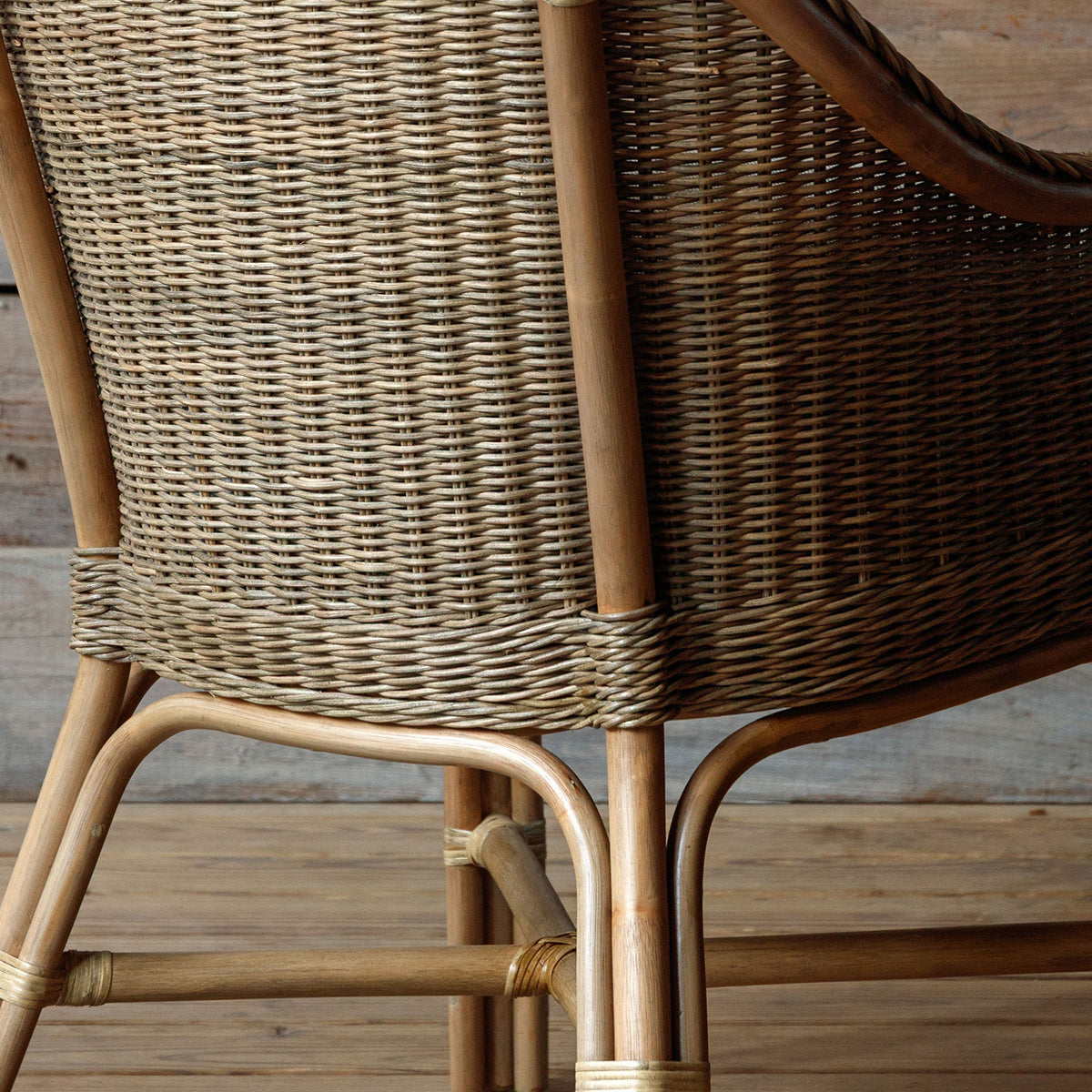 Close-up of a woven rattan chair showing the backrest, armrest, and natural wood frame on a wooden floor.