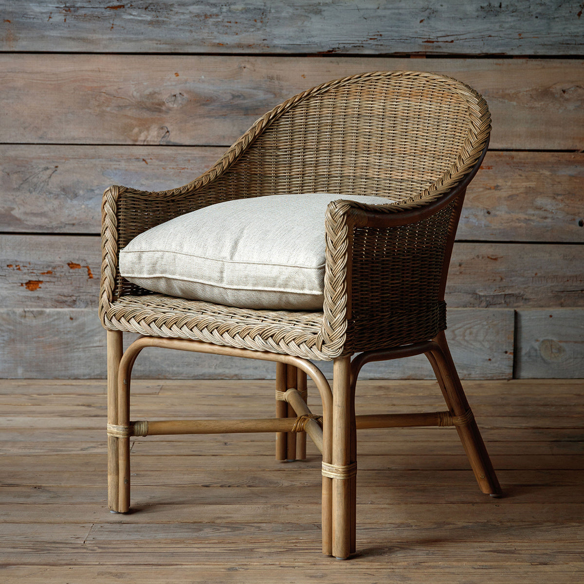 A natural woven rattan armchair with a curved back, rope-wrapped arm details, and a light beige cushion, set against a rustic wood plank wall and floor.