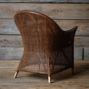 A brown woven rattan armchair with a curved back and light-colored wooden legs, set against a rustic wood plank wall and floor.