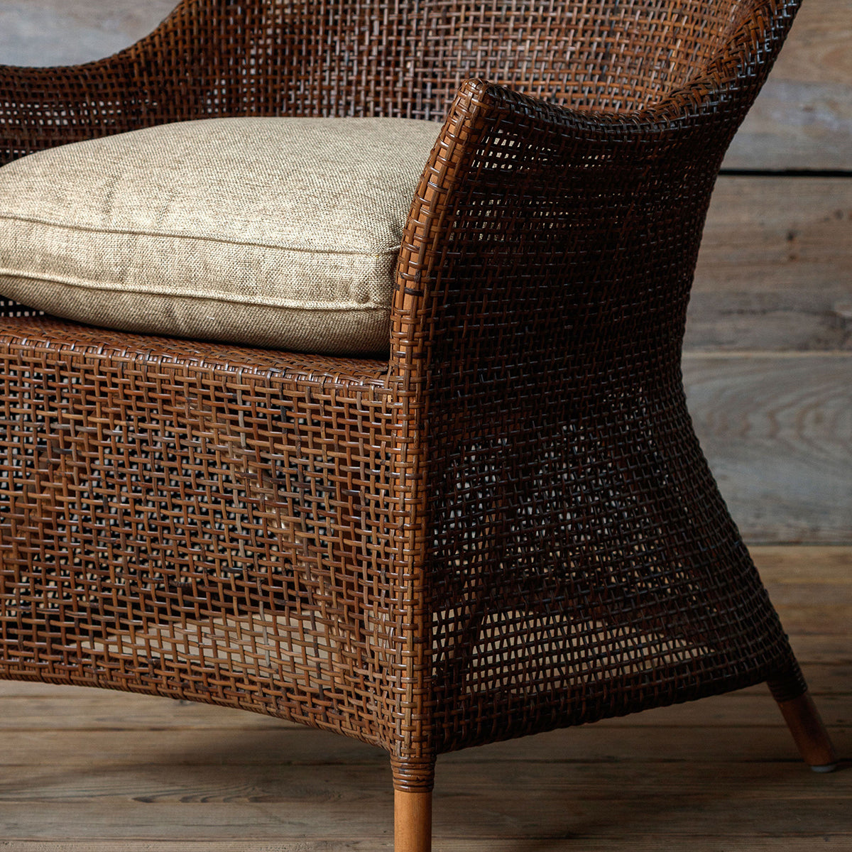 A close-up of a brown woven rattan armchair with a light beige textured cushion on a rustic wooden floor.