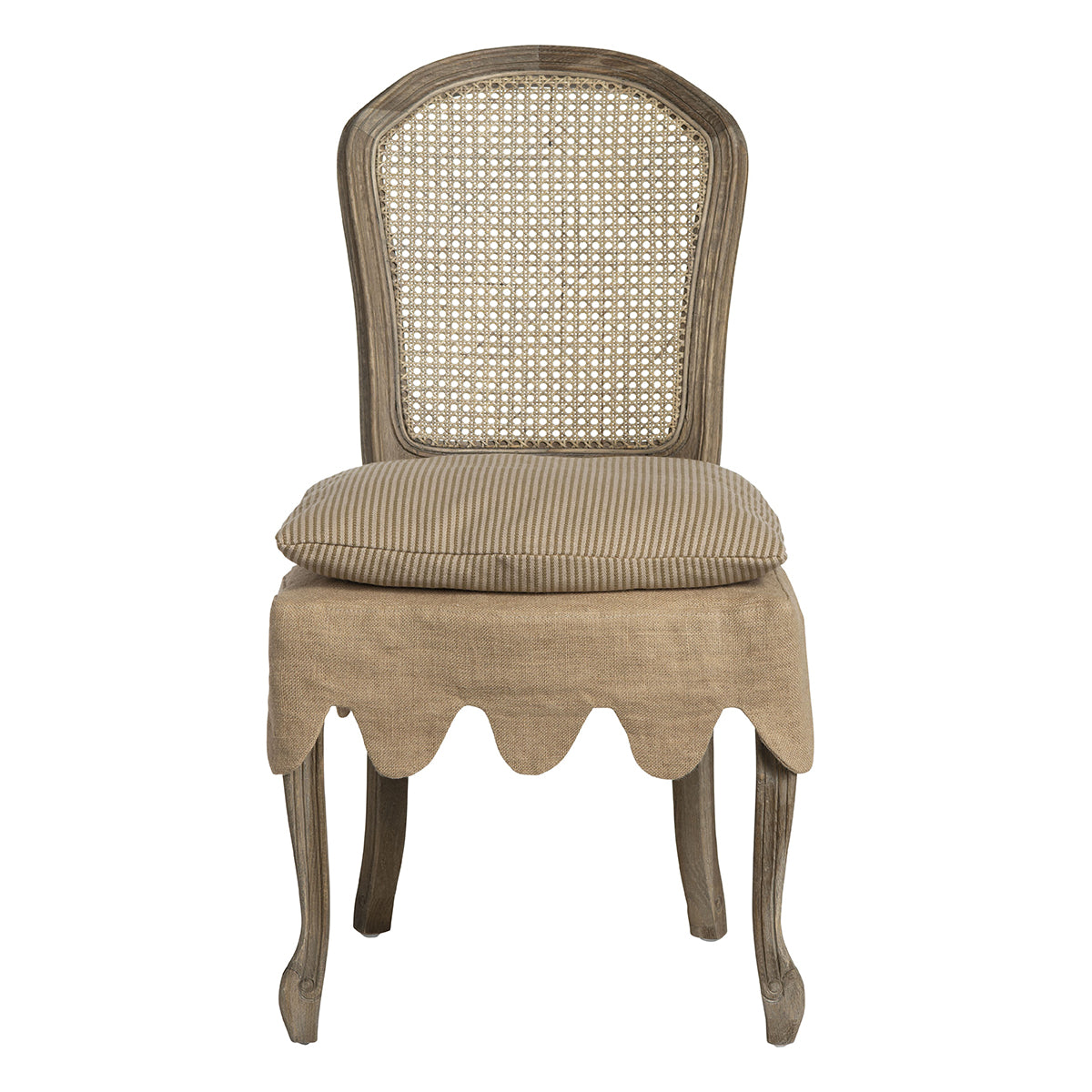 A rustic French-style dining chair with a woven cane backrest, a beige linen cushion, and a scalloped beige fabric skirt over a wooden stool base, set in a vintage room next to a distressed white table.
