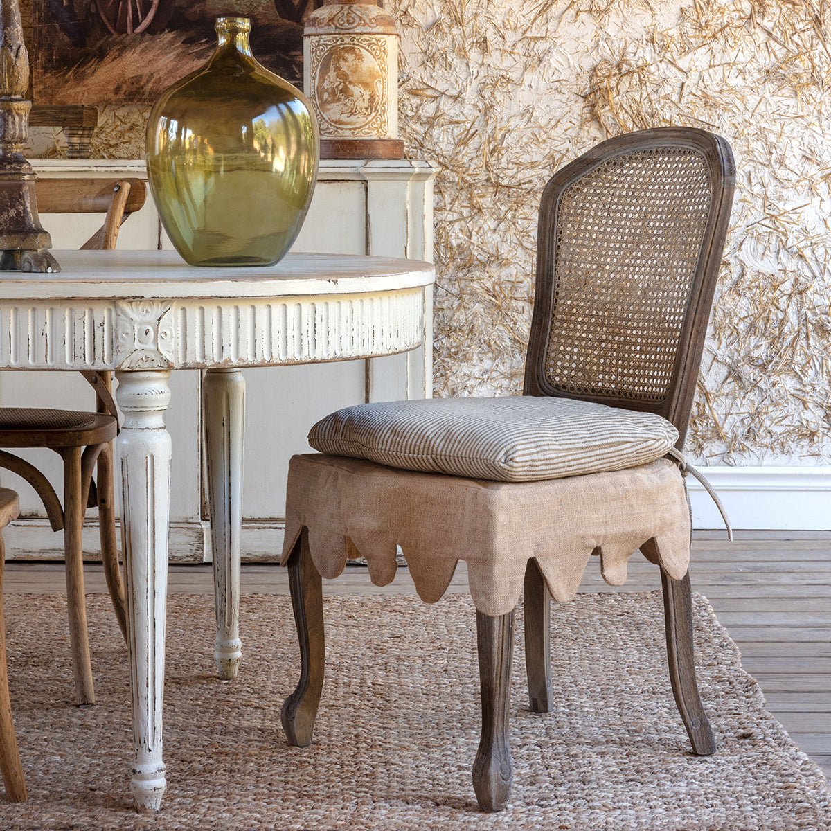 A rustic French-style dining chair with a woven cane backrest, a beige linen cushion, and a scalloped beige fabric skirt over a wooden stool base, set in a vintage room next to a distressed white table.