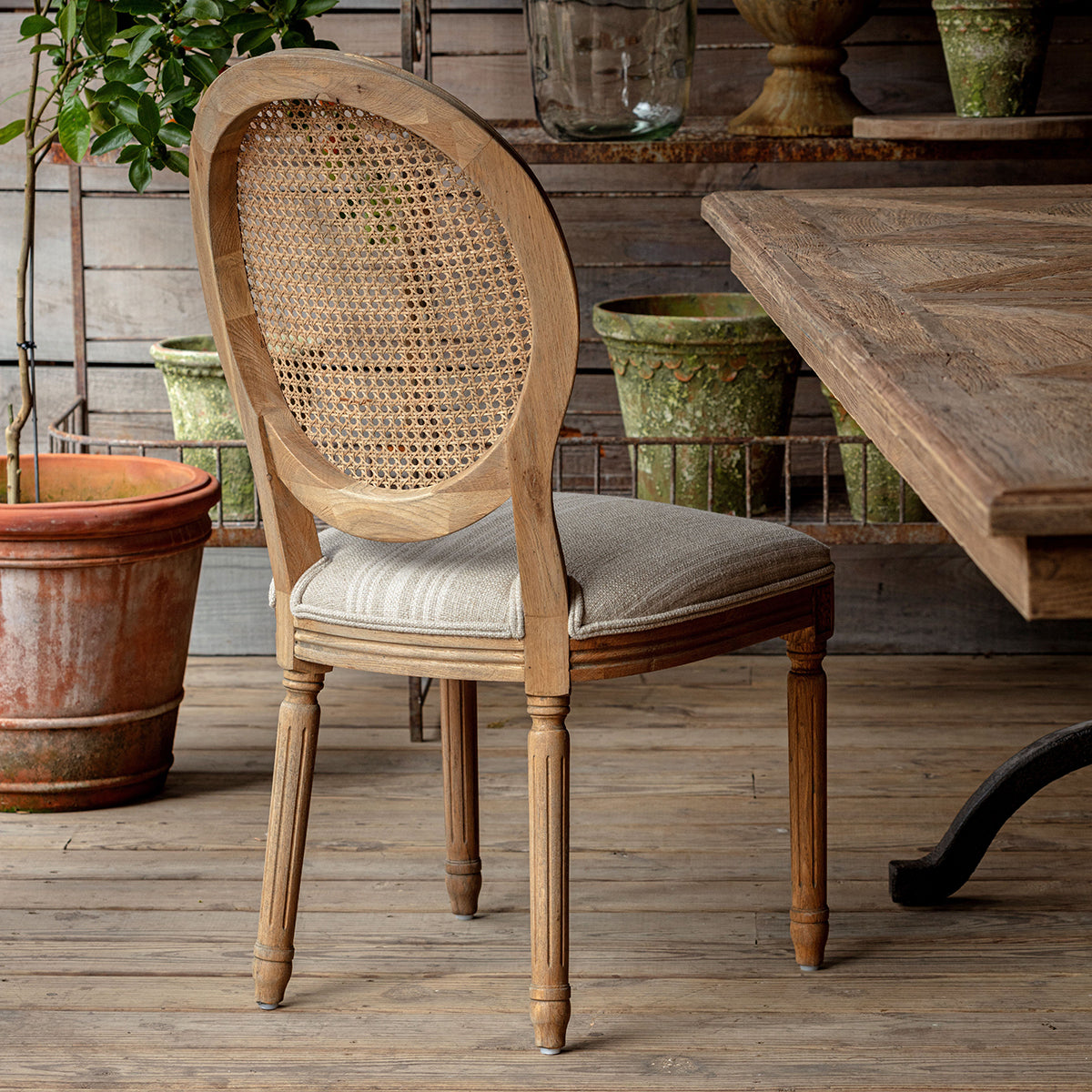 Rosemere Round Cane-Back Dining Chair - Wooden chair with wicker backrest and upholstered seat in a rustic setting with wooden table and potted plants.