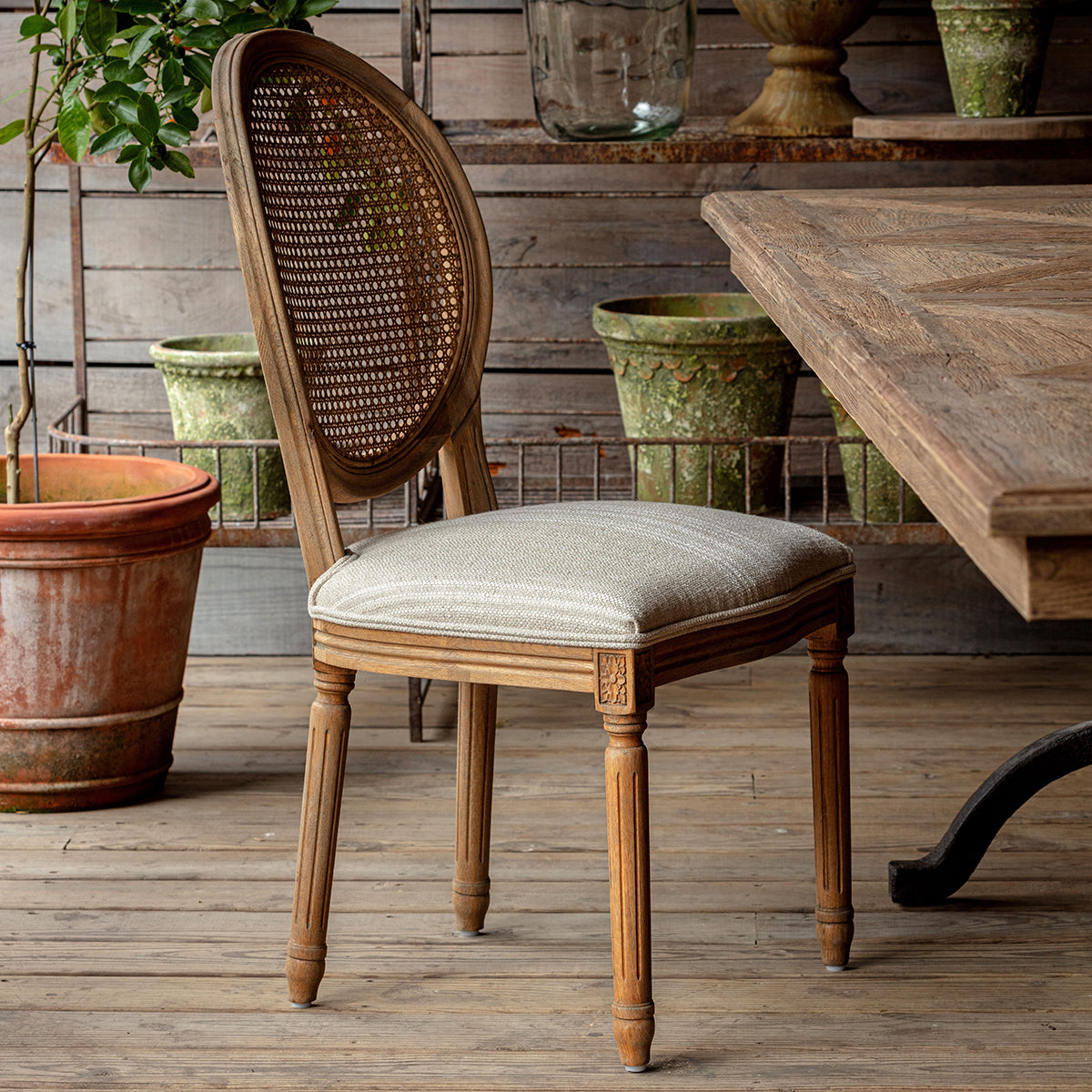 Rosemere Round Cane-Back Dining Chair - Wooden chair with wicker backrest and upholstered seat in a rustic setting with wooden table and potted plants.
