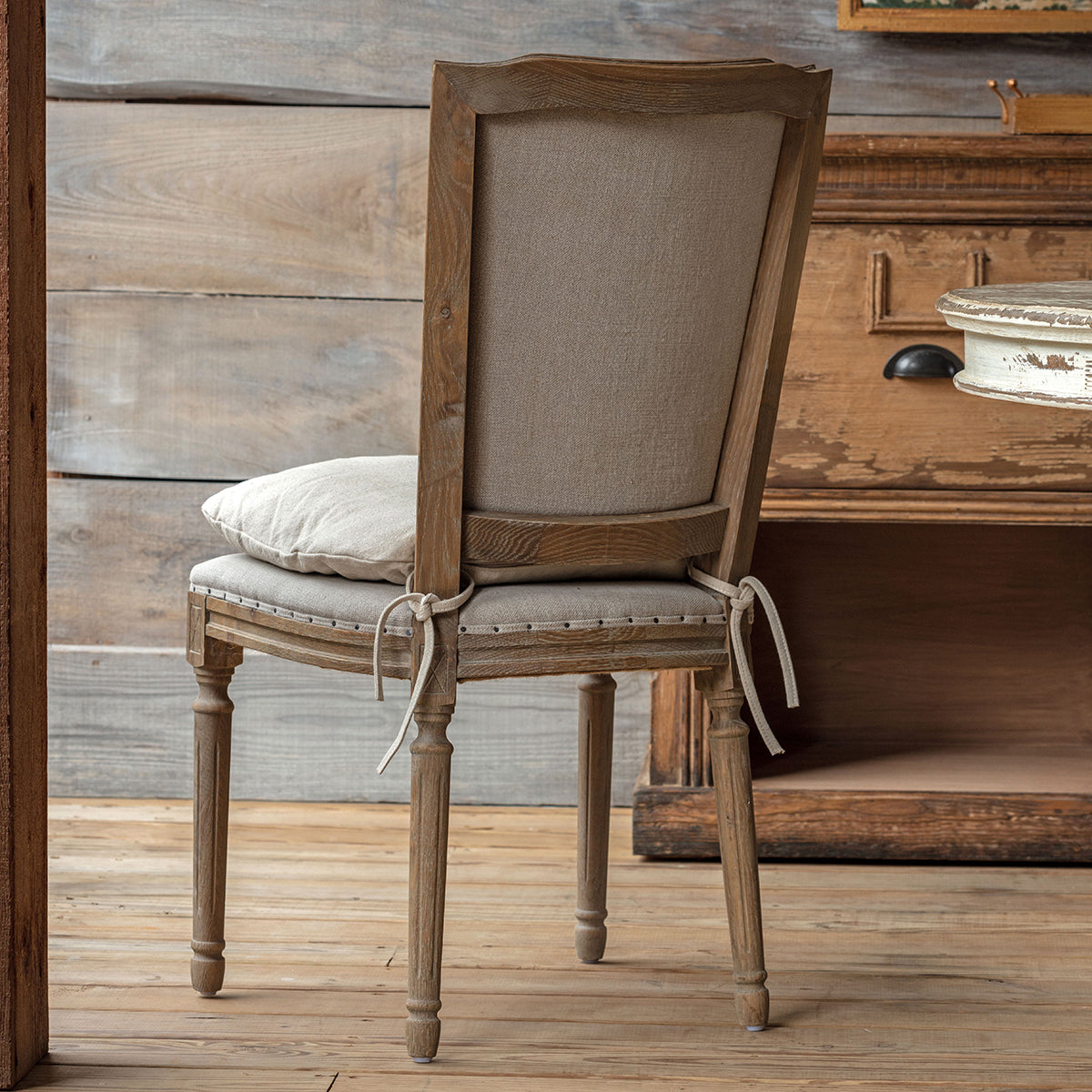 A rustic wooden dining chair with a tall upholstered backrest and a matching cushion, styled in a room with a distressed wooden cabinet and wood plank floor.