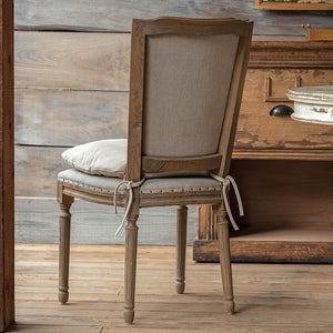 A rustic wooden dining chair with a tall back, upholstered in beige fabric, featuring a matching seat cushion and tie details, set against a distressed wooden background.