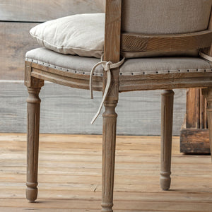 A close-up of a rustic wooden chair with upholstered seat and back in a light beige fabric, featuring a matching cushion and a tied fabric bow on the front.