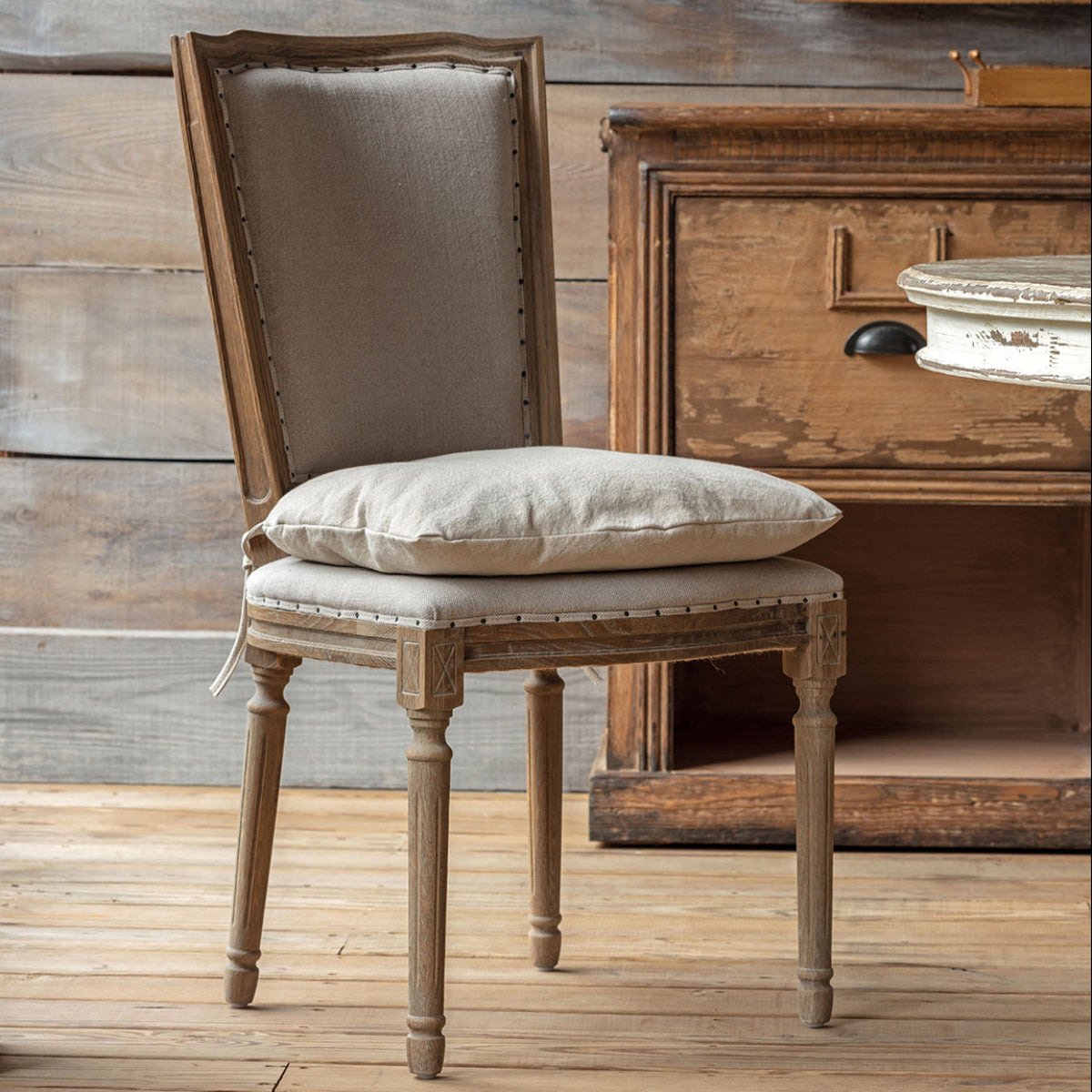 A rustic wooden dining chair with a tall upholstered backrest and a matching cushion, styled in a room with a distressed wooden cabinet and wood plank floor.