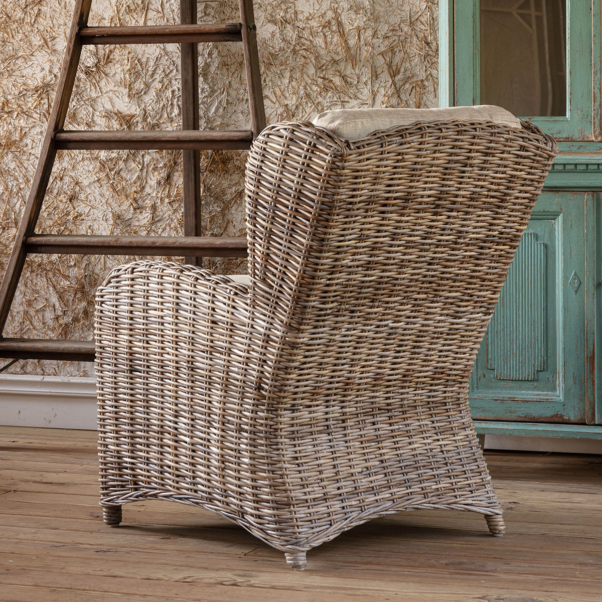 A wicker armchair with a tufted beige back cushion and a light beige seat cushion, set in a rustic room with a wooden ladder, distressed wallpaper, and a teal cabinet.